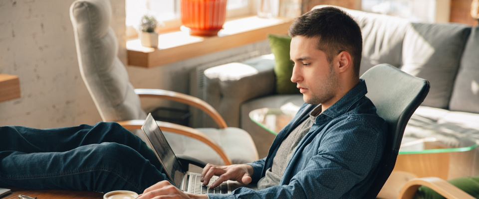 Νεαρός άντρας κάθεται σε πολυθρόνα με το laptop και τον καφέ του. Θεματική εικόνα για συνδυασμό μεταπτυχιακού και δουλειάς.