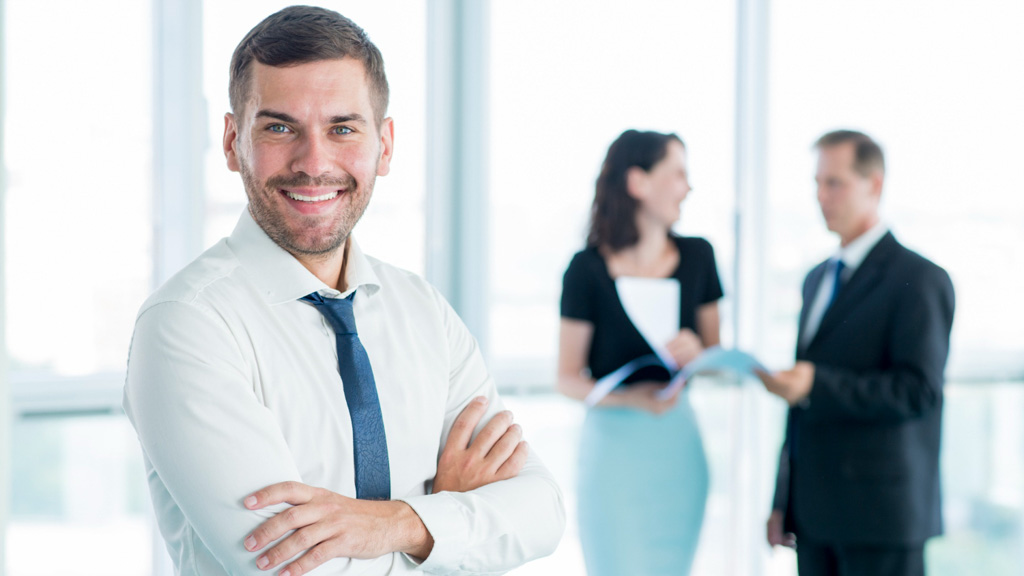 Χαμογελαστός άντρας manager με λευκό πουκάμισο στέκεται μπροστά από δύο συναδέλφους του που μιλάνε μεταξύ τους.
