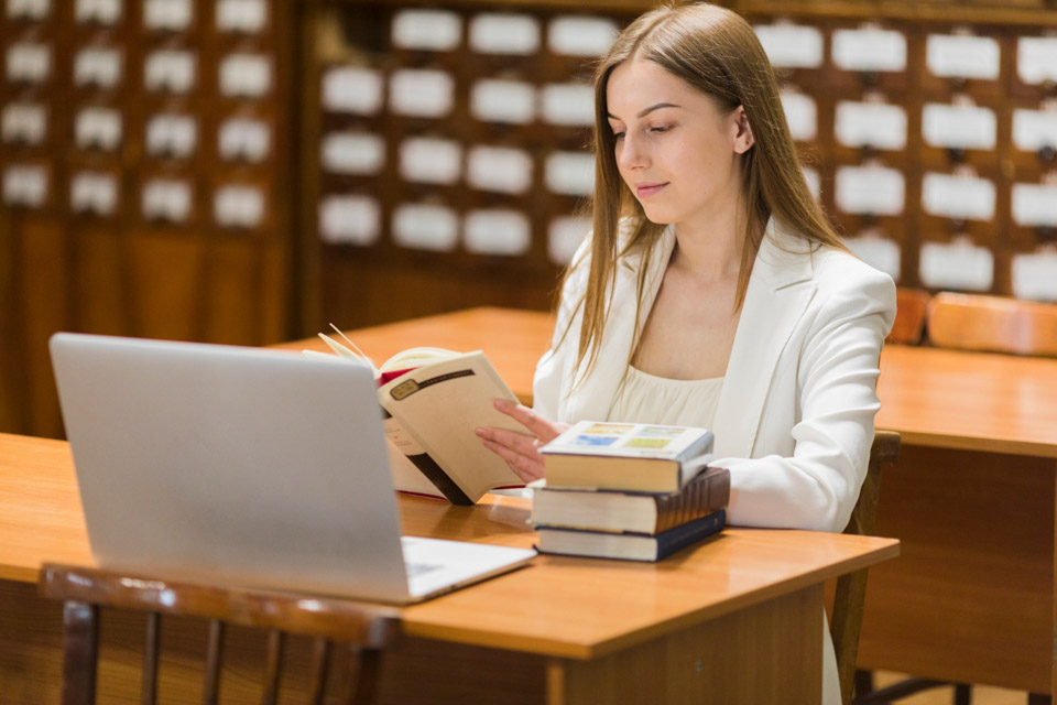 Νεαρή φοιτήτρια κάνει μεταπτυχιακή διπλωματική εργασία, έχοντας ανοιχτό μπροστά της ένα λάπτοπ και διαβάζοντας ένα βιβλίο.